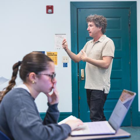 A classroom setting with a teacher standing and speaking. Two students, one focused on papers and the other on a laptop, listen attentively.