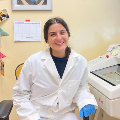 A woman wearing a lab coat and blue gloves smiling in a lab. She is next to scientific equipment.