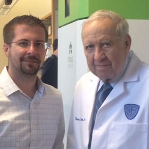 Two men standing side by side, one on left wearing dress shirt and on right white lab coat