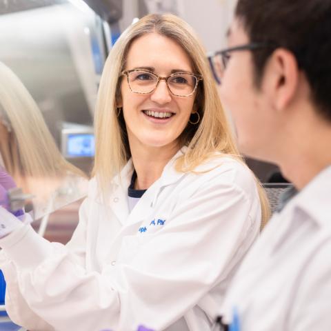 A woman in a lab coat and gloves smiles while holding a test tube rack, engaging with a colleague.