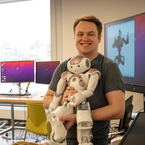 Austin holds a robot in front of a large wall-mounted screen displaying a robot silhouette