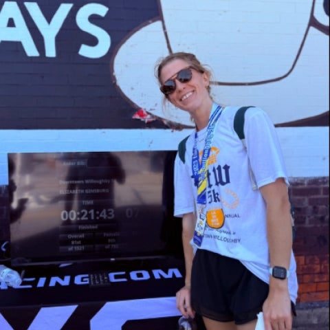 Elizabeth Ginsburg in sunglasses and wearing a race medal stands in front of a computer screen displaying her race time.