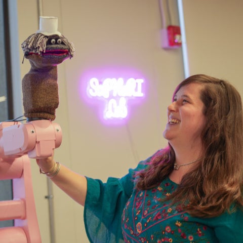 A scientist smiles and looks at a pink robotic arm with a sock puppet