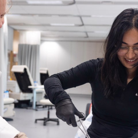 A student in a black shirt practice sutures while a professor in a white lab coat watches and smiles.
