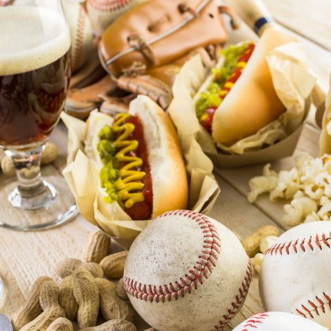 Spread featuring hot dogs, popcorn, peanuts, two glasses of beer, baseballs, a glove, and a bottle opener on a wooden surface.