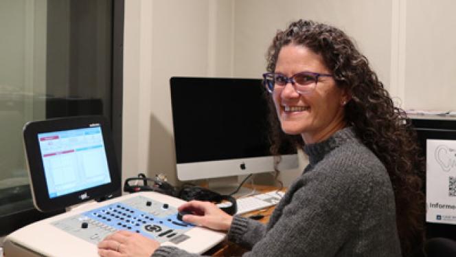 A woman sits with hearing assessment equipment in a sound-isolated booth