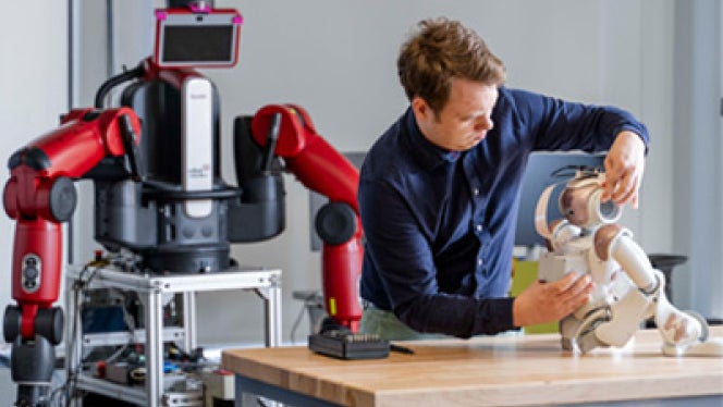 Person stands at a worktable in a robotics lab, adjusting the head of a small white robot seated on the table.