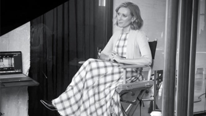 Black and white image of a woman seated in a director's chair, wearing a long checkered dress. She appears calm and focused, with a coffee cup on a side table.