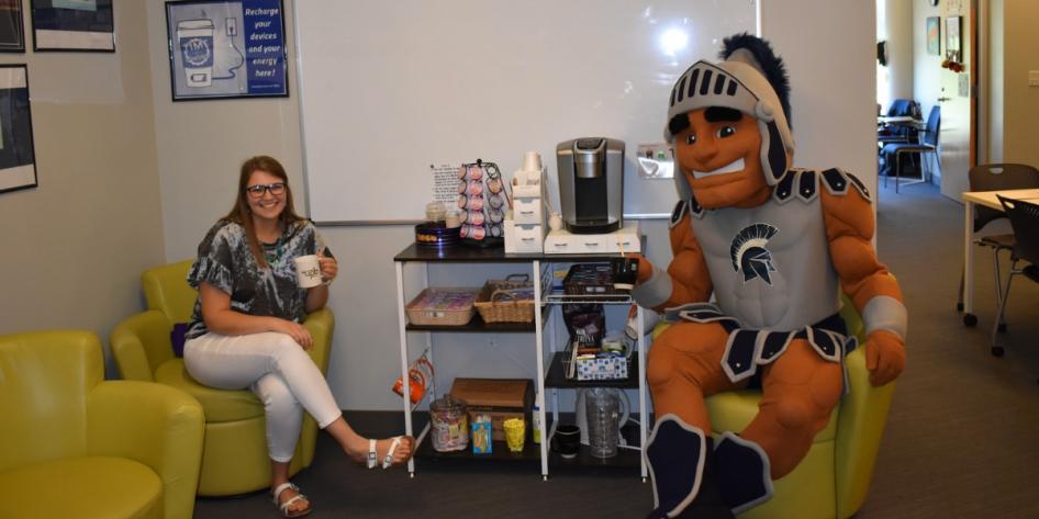 A seated Spartie and staff member holding coffee mugs pose for a photo at a Sippin with Spartie event. 