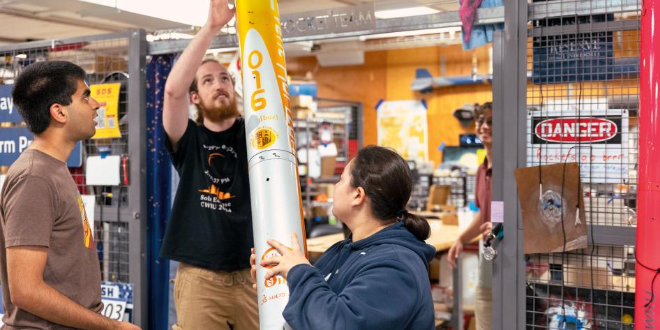 Three people work on a tall white rocket indoors. One lifts the top, another one supports it.