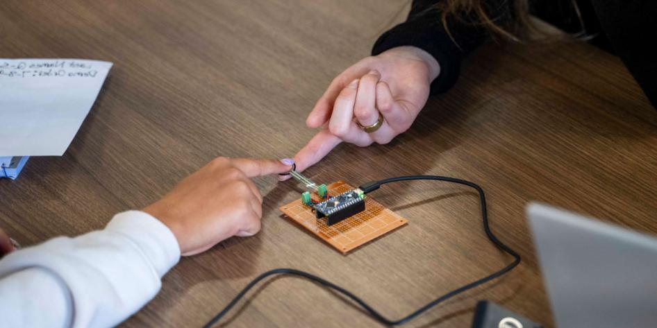 Close up photo of two people using their index fingers to touch a device connected to wires