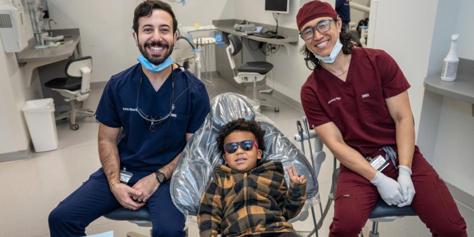 A young boy in a plaid sweatshirt gives a peace sign as he sits in the dental chair between two dental students.