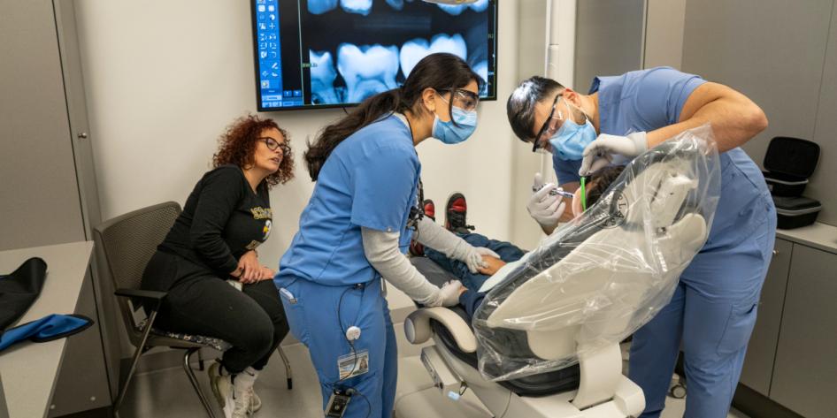 Two dental students perform treatment on a child in the dental chair while his mother observes.