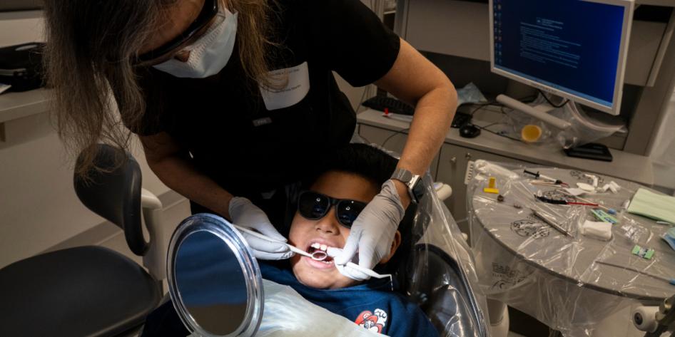A young boy in sunglasses holds a mirror as a dentist examines his teeth.