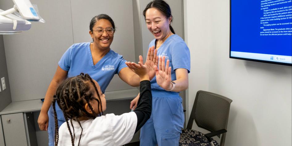 A young girl gives high fives to two dental students in blue scrubs.