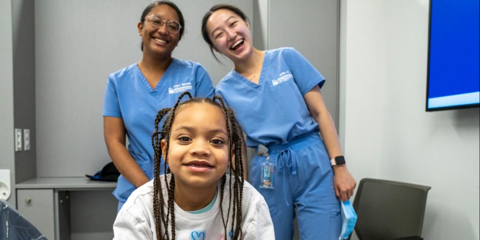 A young girl with braids in a white shirt poses in front of two dental students.