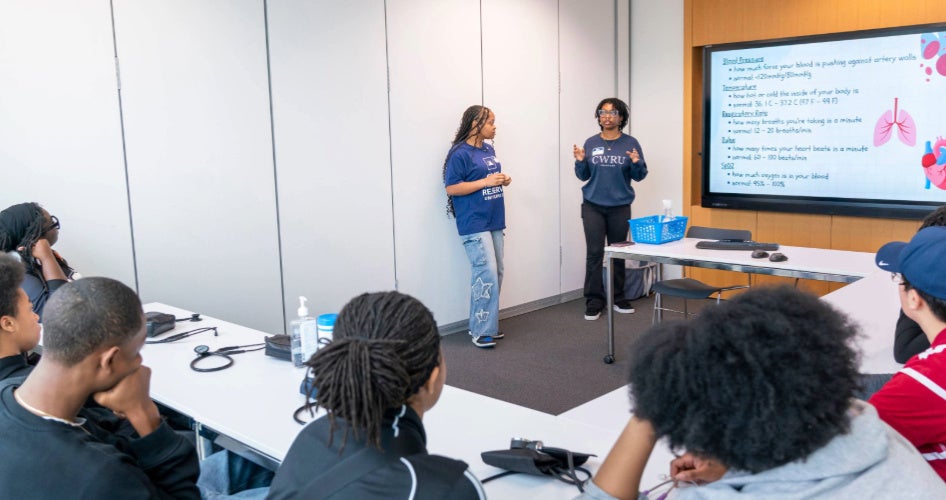 Two nursing students in blue sweatshirts give a presentation to a group of seated students.