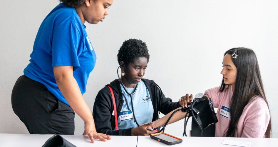 A woman in a blue shirt watches two students practice taking blood pressure.