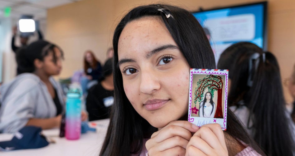 A young woman in glasses holds up a photo of herself.