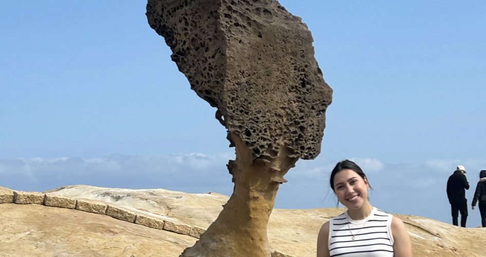 Lopez stands in front of a mushroom rock formation in front of a blue sky and sandy ground.
