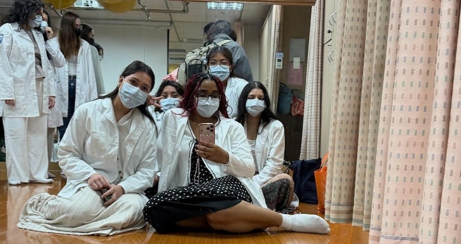 A group of women sits on the floor in white lab coats and medical masks.