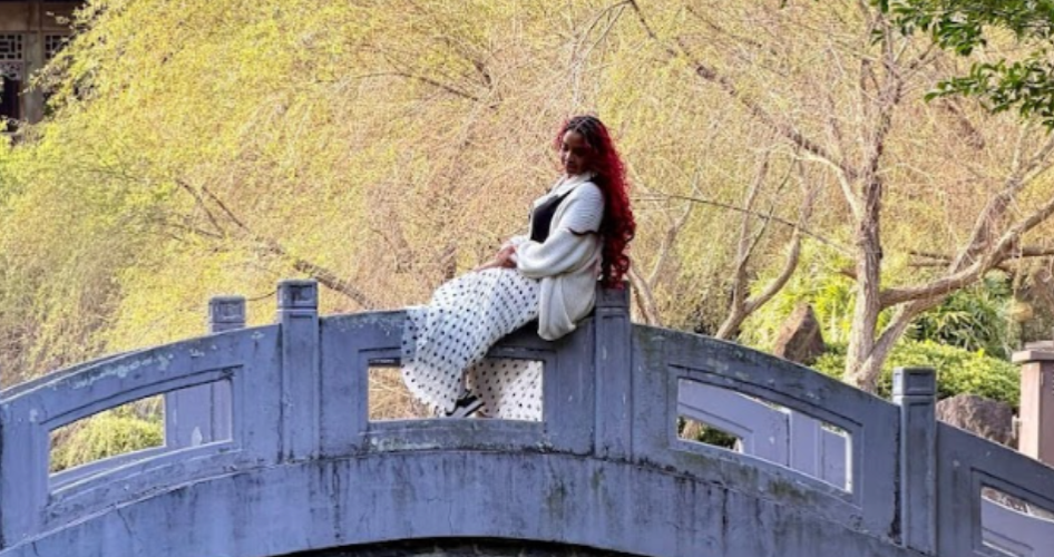 A woman sits on the railing of a stone bridge with trees in the background.
