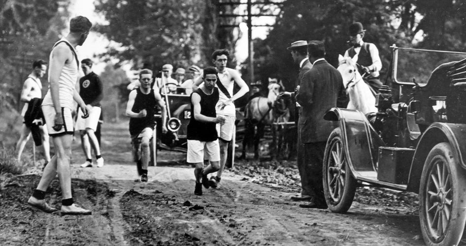 Runners in early 20th-century athletic attire race along a dirt road, with vintage cars and spectators in period clothing lining the path.
