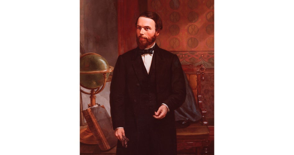 A bearded man in 19th-century attire stands confidently with a globe on a wooden stand beside him and patterned wallpaper in the background. 
