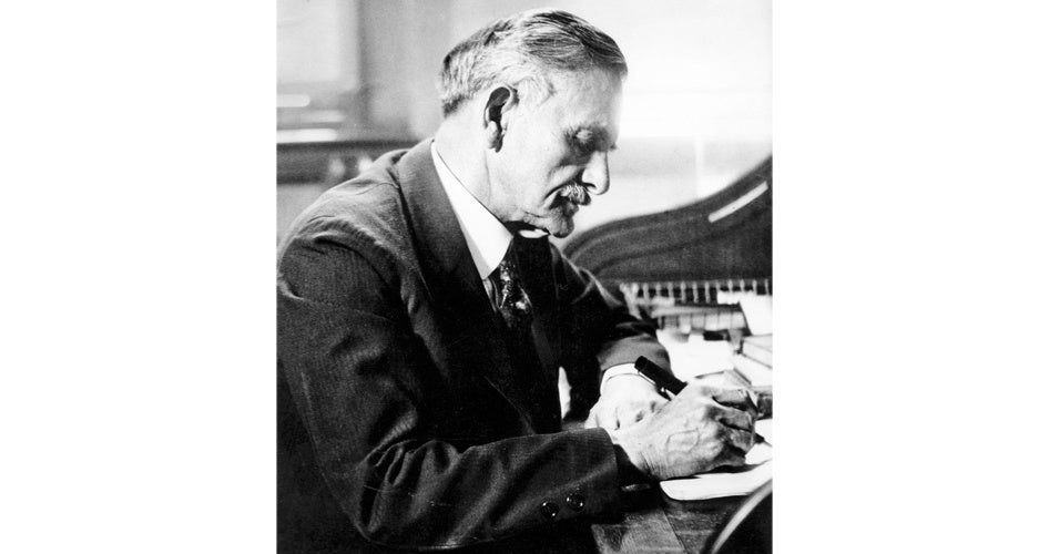 Elderly man in a suit, writing with concentration at a wooden desk. The black-and-white photo conveys a focused, historical atmosphere.