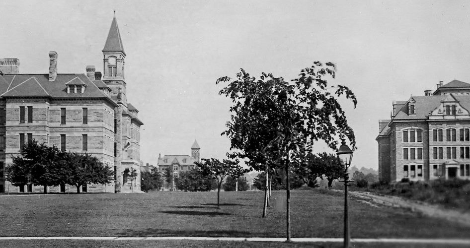 A black and white photo of a university campus with two historic stone buildings featuring towers and pitched roofs, set amidst an open grassy area with scattered trees.