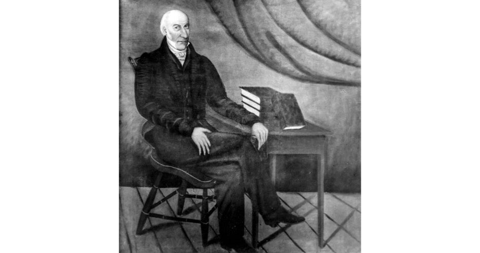 A vintage portrait showing an elderly man in a dark suit sitting on a wooden chair. He is beside a desk with three closed books. The scene is formal.