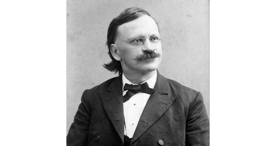 Elderly man with spectacles and a mustache, dressed in a formal suit and bow tie, gazes thoughtfully to the side against a plain background.