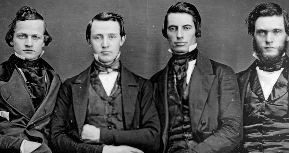 Four young men pose in a vintage black-and-white portrait, dressed in 19th-century suits. They sit closely, with serious expressions, exuding formality.
