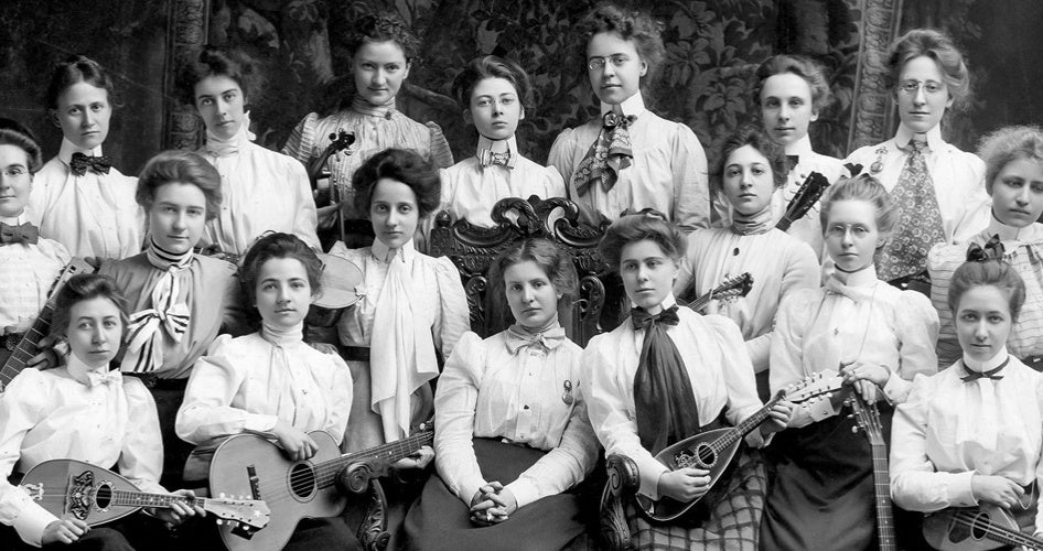 Vintage black-and-white photo of 18 women in Edwardian attire, holding string instruments like guitars and mandolins, exuding a formal and poised atmosphere.
