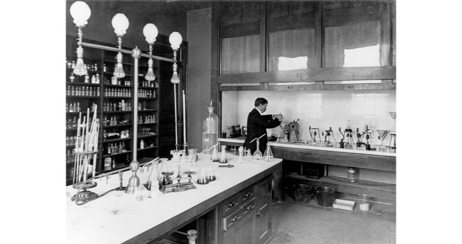A black-and-white photo of a vintage laboratory. A scientist works at a counter filled with glassware, surrounded by shelves of chemicals. The scene appears focused and studious.
