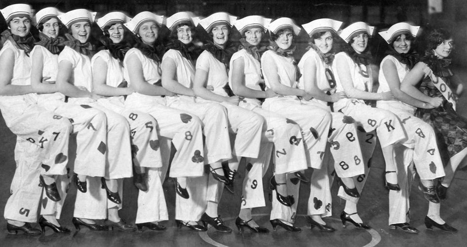 A joyful line of women in sailor hats, wearing outfits decorated with playing card symbols and numbers, pose with wide smiles in a synchronized kick.