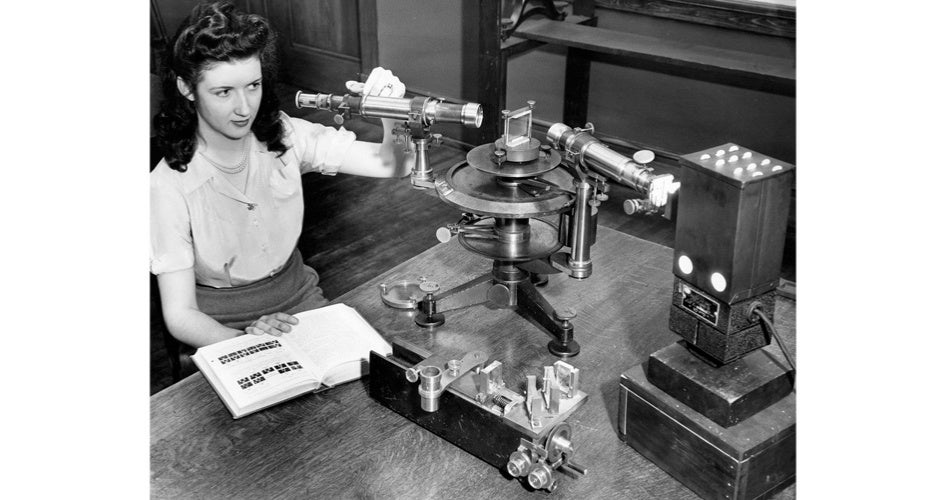 A woman in a vintage laboratory operates a spectrometer, with an open book nearby. The black-and-white photo conveys a sense of focused studying.