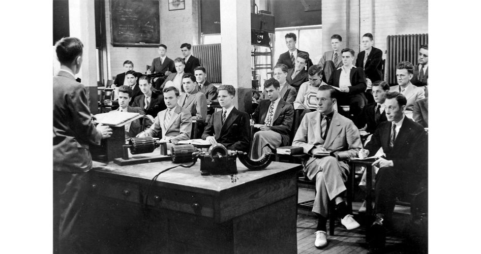 A black-and-white image shows a classroom with men in suits attentively facing a lecturer. The setting is formal, with vintage educational equipment visible.