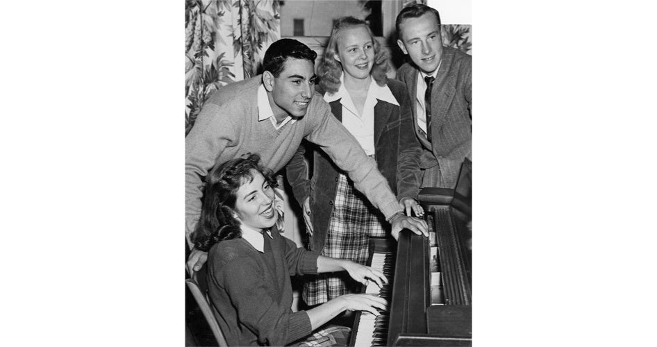 A black-and-white photo of four young adults gathered around a piano. A woman plays joyfully while three others watch, smiling. The scene is lively and warm.