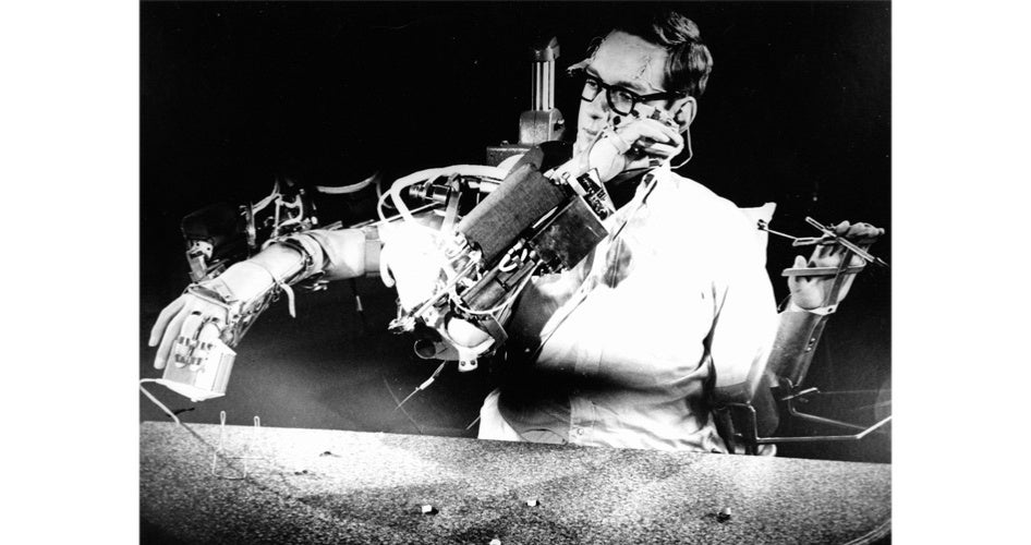 Black and white photo of a man operating a robot with mechanical arms, holding a cup and a tool. The scene conveys innovation and focus.