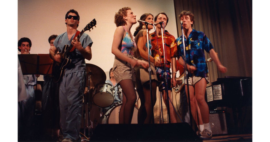 A lively band performs on stage. Musicians are dressed in colorful, casual attire, playing instruments and singing into microphones, exuding energetic vibes.