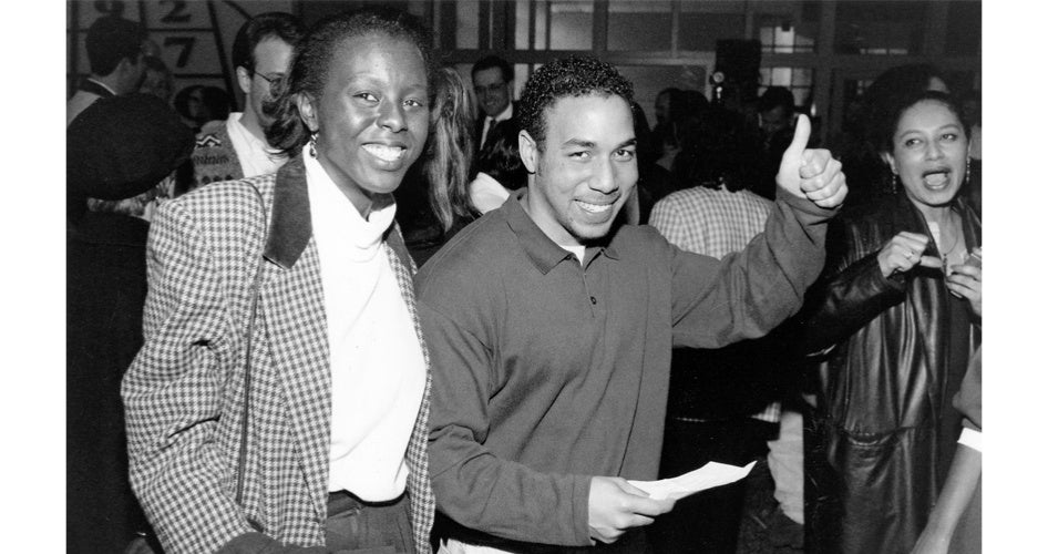 A smiling man holds a paper and gives a thumbs-up gesture, standing beside a woman in a checkered coat. The background shows a lively crowd.
