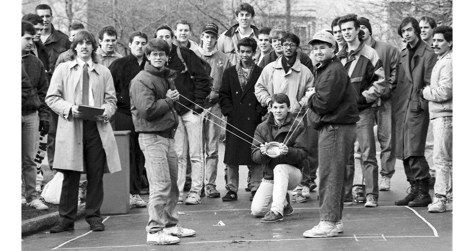 A group of young men stand outdoors, centered around a homemade catapult, exuding teamwork and enthusiasm. The atmosphere is lively and collaborative.