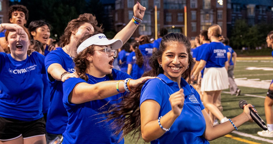 A group of young people in blue shirts joyfully celebrate on a sports field. The mood is energetic and festive, with smiles and raised arms.