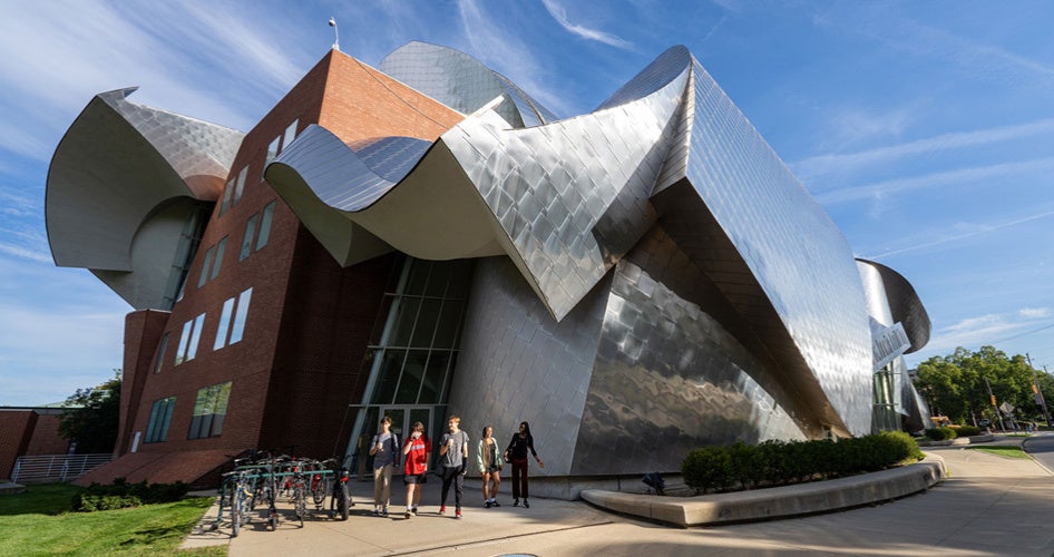 A uniquely designed modern building with abstract, curved silver panels and red brick walls under a blue sky. Five people walk in front.