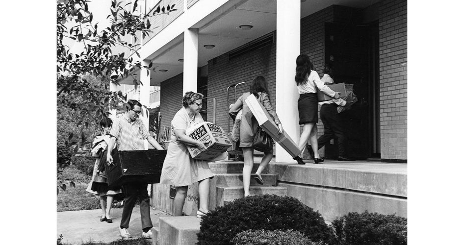 A group of people carrying boxes and suitcases enters a building, suggesting moving in or out. The scene is lively and collaborative. Black and white.