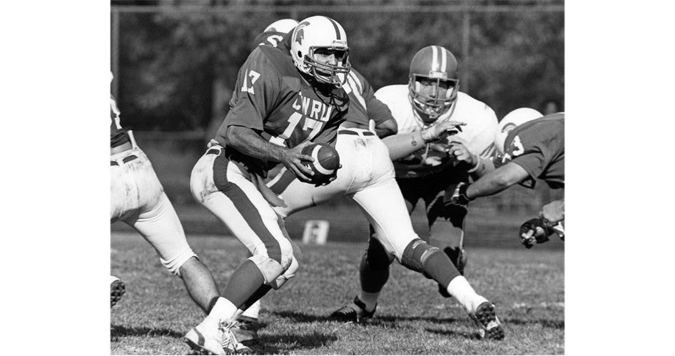Black and white image of a football quarterback in a "CWRU" jersey, number 17, holding the ball, surrounded by players in action on a grassy field.