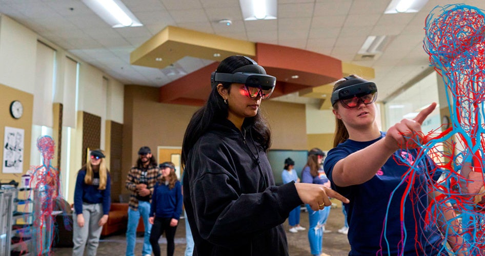 Two people wearing VR headsets interact with a digital, anatomical human model in a classroom. Others in the background also use headsets.