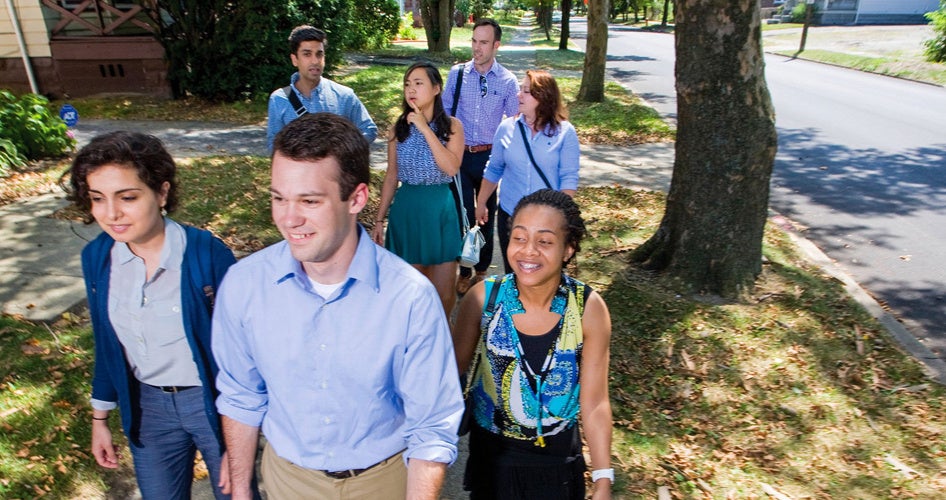 A diverse group of people smiling and walking on a tree-lined sidewalk in a suburban neighborhood, conveying a sense of camaraderie and happiness.