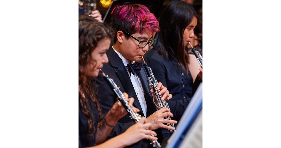 A focused group of oboe players in a formal orchestra, each intensely engaged in reading sheet music. The scene conveys concentration and musical collaboration.